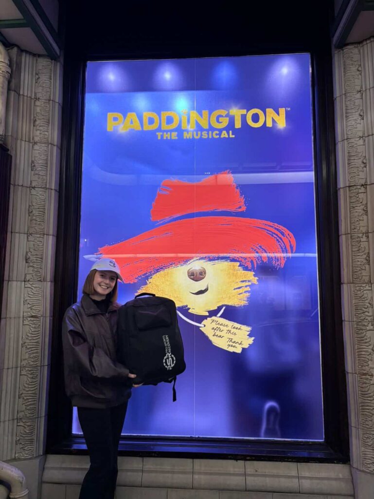 Rose Mary O'Reilly holding the Pro Sports Recovery Normatec bag in front of a Paddington the Musical poster