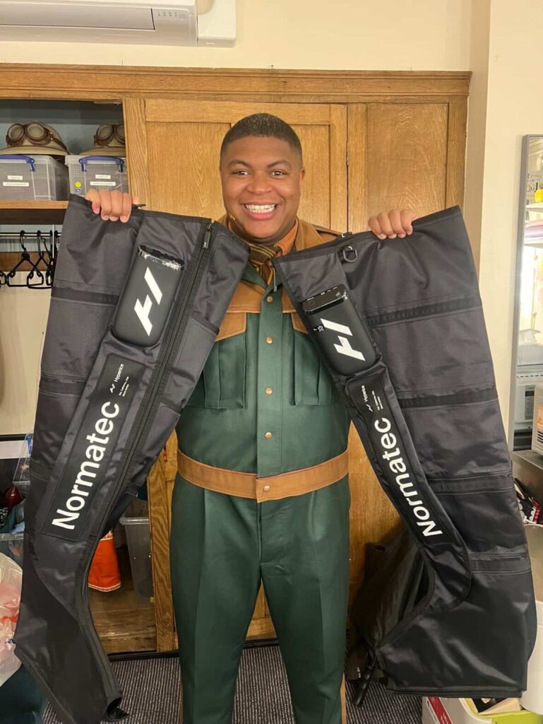 Tarinn Callender holding open Normatec compression boots backstage at Paddington the Musical 